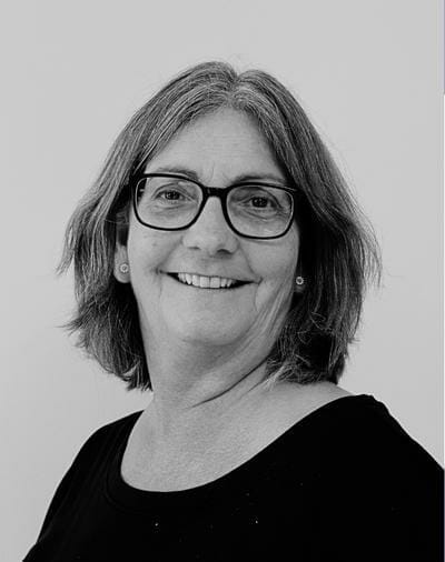 Et gråtoneportræt af en smilende kvinde med skulderlangt hår og briller. Hun er iført en sort top og små øreringe, mod en almindelig baggrund.