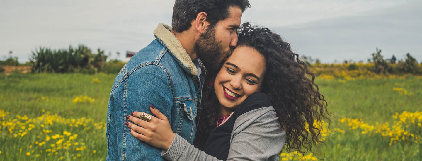 A man wearing a denim jacket kisses the forehead of a smiling woman with curly hair. They are in a field with green grass and yellow flowers, under a cloudy sky.