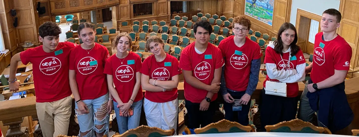 A group of eight people wearing matching red shirts with a logo stands inside a large hall with wooden interiors and rows of green chairs. They appear relaxed and are gathered closely, some smiling at the camera.