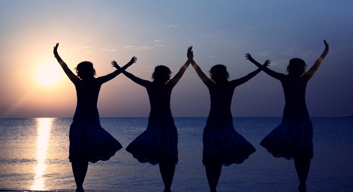 Silhuetter af fire personer med hævede arme, stående i en linje ved kysten ved solnedgang. Solen står lavt i horisonten og kaster et varmt skær over det stille hav og himmel.