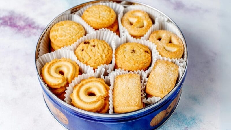 Blå blikæske fyldt med et udvalg af smørkager i hvide papirkopper. Småkagerne har forskellige former, herunder hvirvler, kringler og rektangler, hver let gyldenbrune på toppen. Dåsen er på en marmoreret overflade.