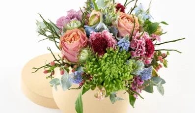 A round beige box filled with an arrangement of pink roses, green chrysanthemums, small purple flowers, and assorted greenery on a white background. The flowers overflow, adding a lush, colorful touch to the composition.