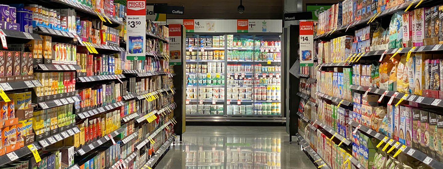 En købmandsgang fyldt med forskellige kornkasser på begge sider. I baggrunden er der kølehylder fyldt med mælk og drikkevarer. Prisskilte og kampagner er synlige på hylderne.