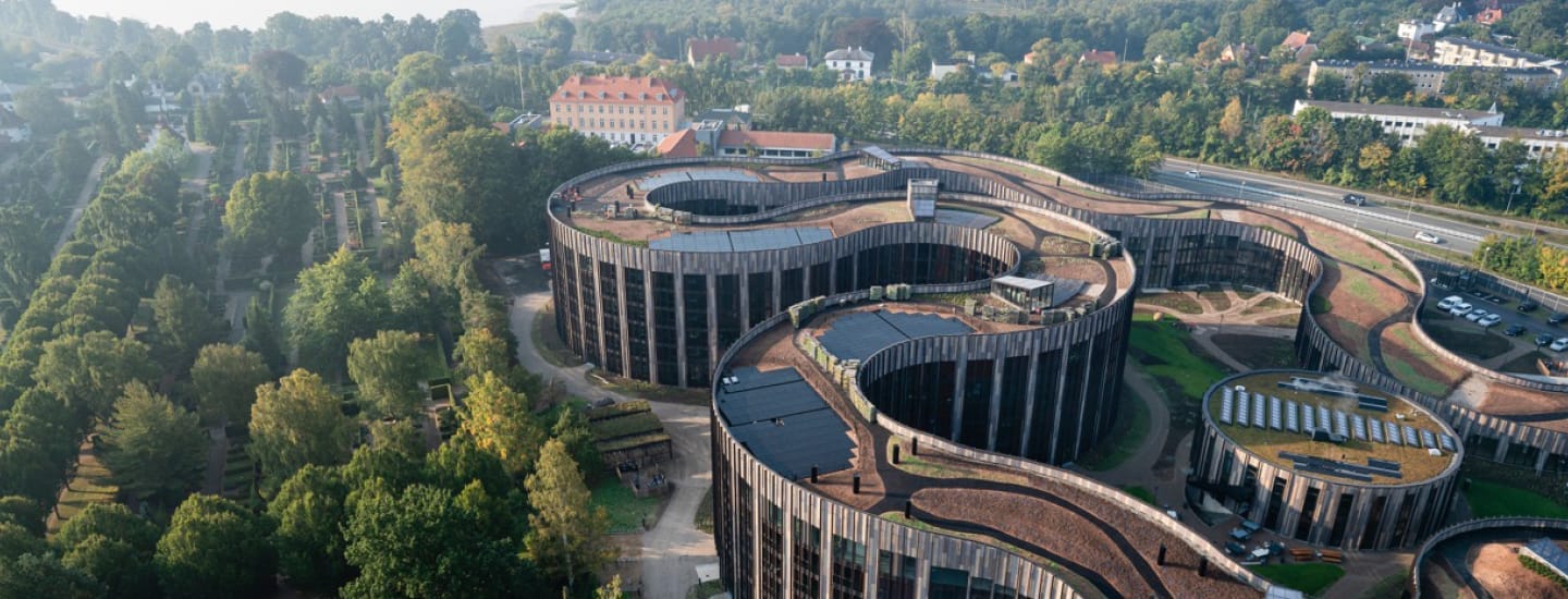 Luftfoto af en moderne, snoet, cylindrisk bygning med græsklædte tage omgivet af frodige grønne områder og træer. I nærheden ligger veje og boligområder med huse synlige i det fjerne.