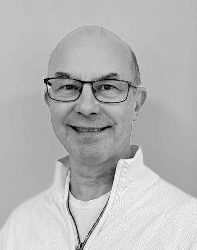 En smilende mand med briller og skaldet hoved, iført en hvid trøje med lynlås, poserer mod en almindelig baggrund på et sort/hvidt foto.