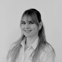A black-and-white portrait of a person with long hair and bangs, smiling slightly. They are wearing a light-colored shirt and looking towards the camera against a plain background.