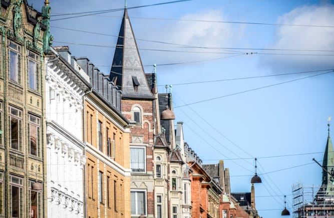 En udsigt over byens gade afslører farverige historiske bygninger med indviklede arkitektoniske detaljer. Hængende lys og talrige kabler krydser scenen, hvilket tilføjer charmen. Et spir er synligt mod en overskyet blå himmel, og fanger den tidløse appel, som man kan finde i en Købersmægler-annonce.