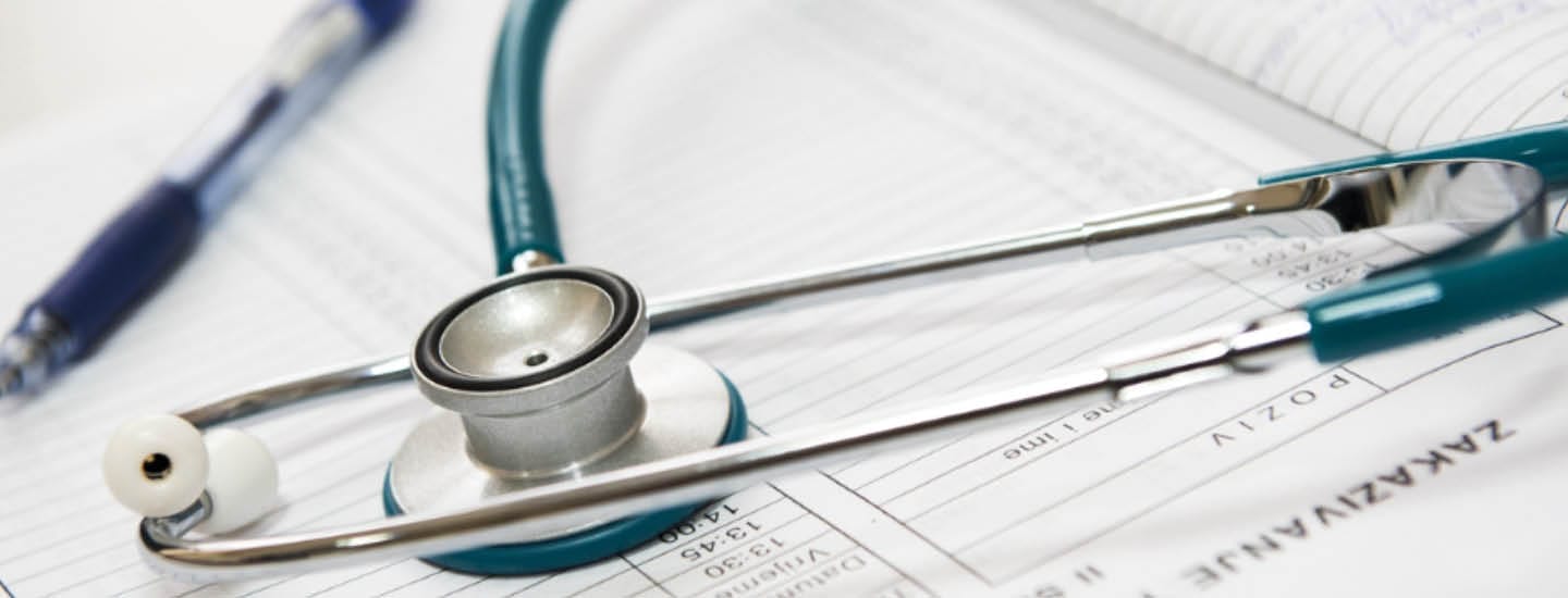 Et stetoskop hviler på et åbent medicinsk dokument ved siden af en pen. Stetoskopets ørestykker og bryststykke er synlige, hvilket understreger en sundhedsmæssig indstilling. Dokumentet indeholder tabeller og tekst, der angiver lægejournaler eller data.