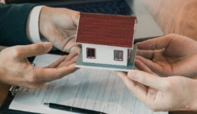 Two people in business attire hold a small house model over a document on a table. A pen and a laptop are partially visible, indicating a real estate transaction or discussion.
