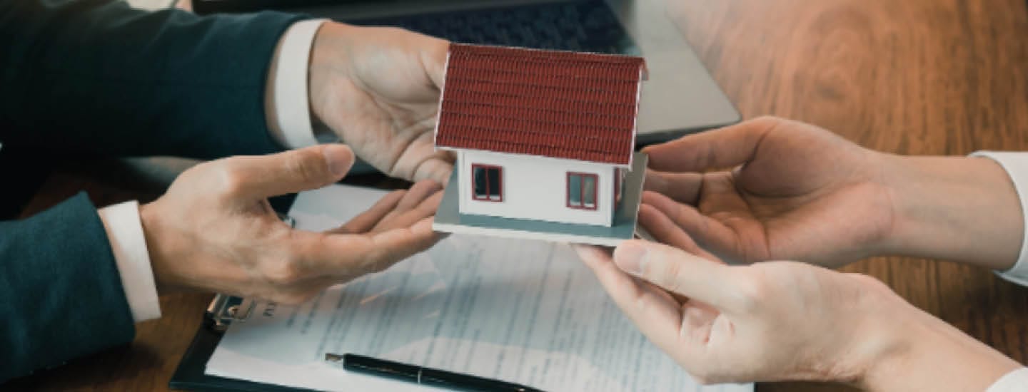 To personer, der holder et lille modelhus over en udklipsholder med papir, kuglepen og en bærbar computer på bordet illustrerer den minutiøse proces i en ejendomshandel, muligvis styret af en Købersmægler.