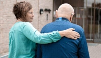 A woman in a teal shirt lovingly guides an elderly man in a blue sweater, who uses a hearing aid. They walk side by side outdoors, near a brick building with large glass doors.