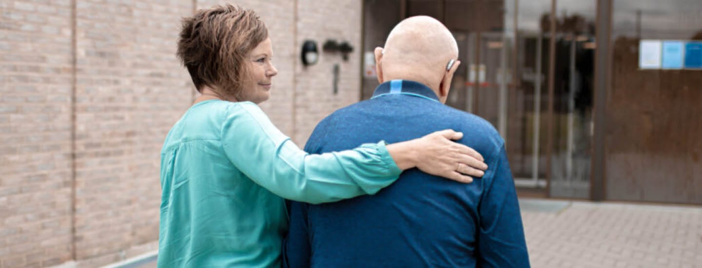 En kvinde i en blågrøn bluse støtter en ældre mand med et høreapparat, der går ved siden af ham. De er uden for en bygning med murstensvægge og glasdøre. Kvinden har armen om mandens skulder og giver omsorg og kammeratskab.