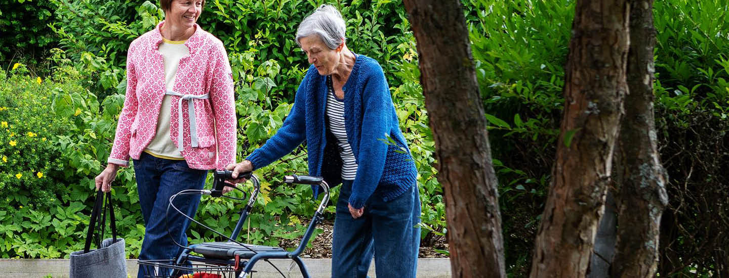To ældre kvinder går i en have. Man bruger rollator og har en blå cardigan på; den anden har en grå mulepose og har en pink cardigan på. De er på en asfalteret sti, omgivet af frodigt grønt løv og træer.