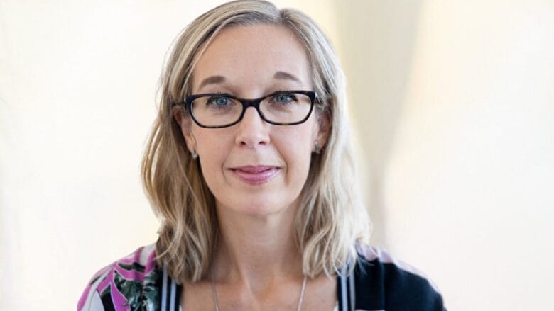 En person med skulderlangt blondt hår og briller ser på kameraet med et neutralt udtryk. De har en mønstret top og en halskæde på. Baggrunden er blødt sløret.