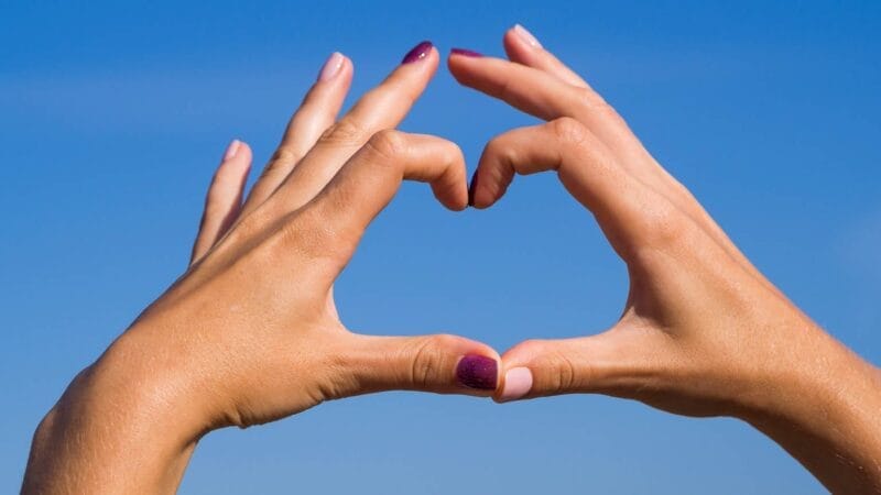 To hænder danner en hjerteform mod en klar blå himmel. Fingrene er låst sammen ved spidserne, hvilket skaber omridset af et hjerte. Den ene hånd har lilla neglelak, mens den anden har lyserød.
