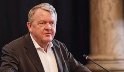 En mand i jakkesæt taler ved en mikrofon foran en sløret baggrund med en søjle.