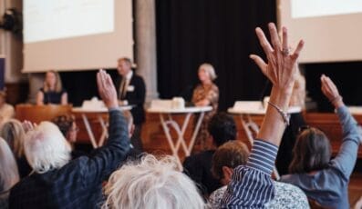 Folk i et publikum, der rækker hænderne op på en konference eller et seminar med talere stående ved et panel i baggrunden. Rammerne er indendørs med fokus på engagement og interaktion.