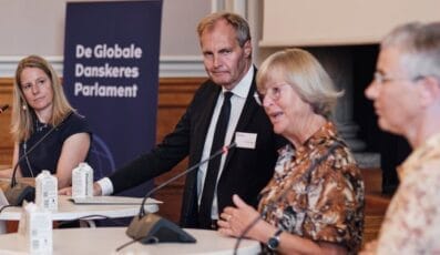 Fire personer er engageret i en diskussion ved et konferencepanel. Tre sidder, en står. På et skilt i baggrunden står "De Globale Danskeres Parlement." De har mikrofoner og sidder ved borde med drikkevarekartoner.