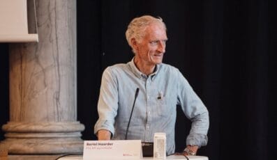 Ældre mand med hvidt hår står ved et podium med en mikrofon iført en lyseblå skjorte. Et navneskilt er synligt på podiet. En stensøjle og et mørkt gardin er i baggrunden.