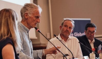 Ældre mand taler i en mikrofon ved en paneldebat, med tre personer siddende ved siden af ham. De er i et mødelokale med et projektorlærred bag sig.