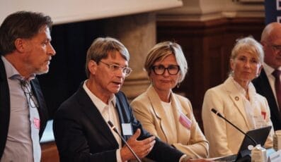 En gruppe på fem personer i formel påklædning står ved et podium. Mellempersonen taler, mens de andre lytter opmærksomt. Der er papirer og mikrofoner på podiet, og en stor skærm er delvist synlig i baggrunden.