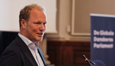 En person i et jakkesæt taler til en konference og står ved siden af en mikrofon. I baggrunden vises et blåt banner med delvist synlig tekst.