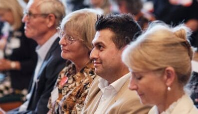 En gruppe mennesker, der sidder på en række. En mand i hvid skjorte sidder mellem to kvinder, den ene iført briller og en mønstret skjorte, den anden i beige outfit. Flere mennesker sidder i baggrunden.