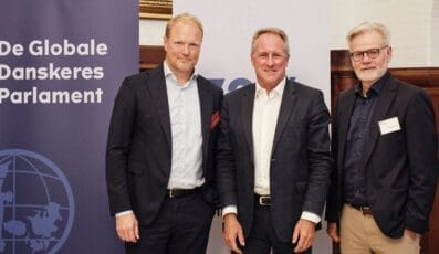 Tre mænd i jakkesæt står smilende foran bannere med tekst, hvor der står "De Globale Danskeres Parlament" og en globusgrafik. Baggrunden foreslår en indendørs begivenhed eller konference.