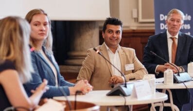 En paneldebat med fire personer siddende ved et bord med mikrofoner. Manden i midten smiler, iført en solbrun blazer. To kvinder til venstre lytter opmærksomt, og en anden mand sidder til højre i et jakkesæt.