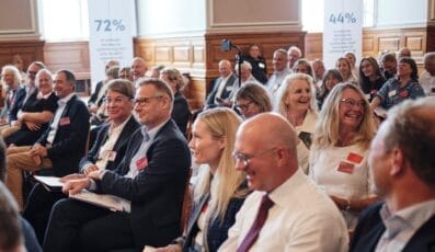 Folk sidder i et mødelokale og deltager i en session. Nogle lytter opmærksomt, mens andre smiler og taler. To plakater i baggrunden viser procenter og tekst, og træpaneler pryder væggene.