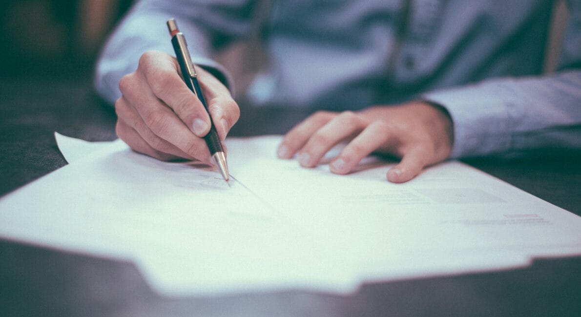 En person i en blå skjorte skriver på en stak papirer med en kuglepen, siddende ved et mørkt skrivebord. Fokus er på hænderne, med baggrunden lidt sløret, hvilket tyder på et arbejds- eller studiemiljø.
