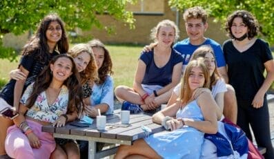 En gruppe smilende unge mennesker, måske en del af Danes Worldwide, sidder og står omkring et picnicbord udendørs. Nogle sidder på bordet, mens andre slapper af på bænken eller står. Solrigt vejr bader dem i varme, med grønne træer, der giver en fredfyldt kulisse.