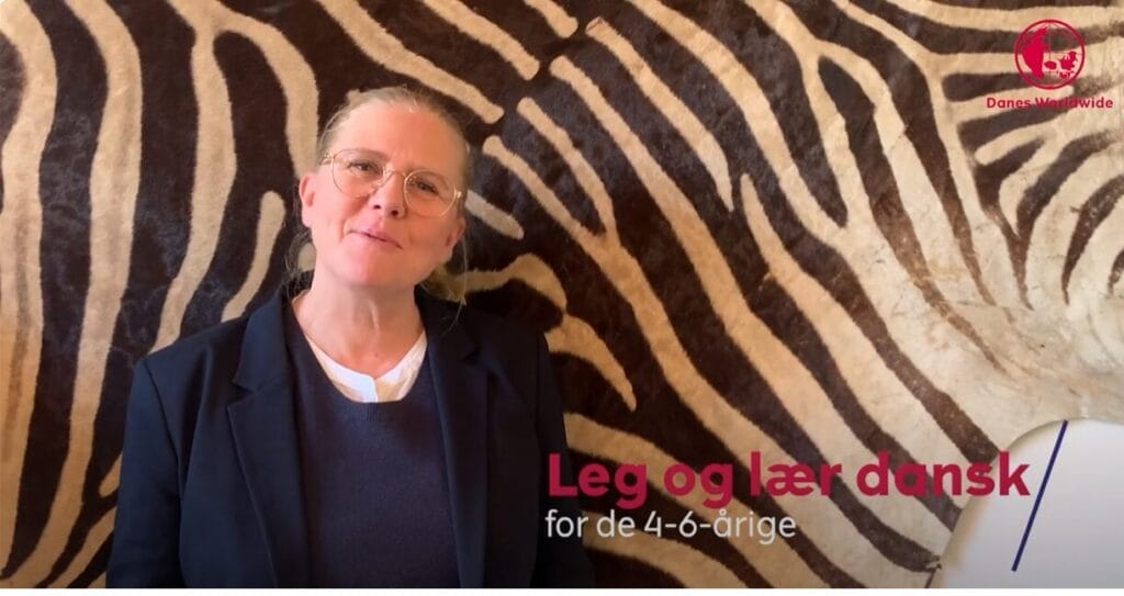 A person wearing glasses and a black jacket stands in front of a zebra-patterned background. Text on the image reads, Leg og lær dansk for de 4-6-årige, with the Danes Worldwide logo in the corner.