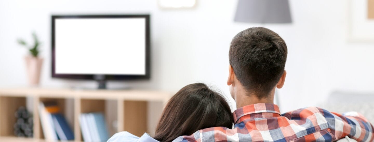 Et par sidder i en sofa og ser fjernsyn med ryggen mod kameraet i et blødt oplyst rum udsmykket med Bloomit-indretning. En hylde rummer bøger og dekorative genstande, der kaster blide skygger, mens en lampe ved siden af dem tilføjer varme. Personen til venstre hviler hovedet på den andens skulder.