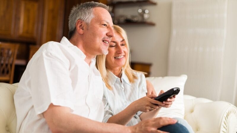 A middle-aged couple sits on a white couch in a cozy living room. The man, wearing a white shirt, and the woman, holding a remote control, are smiling and looking in the same direction. Wooden furniture is visible in the background.