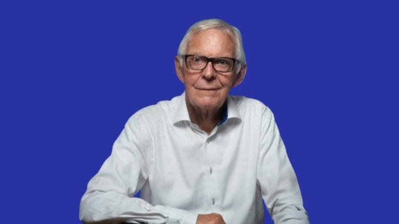 Ældre mand med briller og hvidt hår iført hvid skjorte, siddende mod en solid blå baggrund.