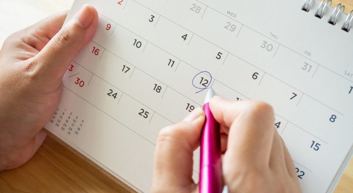 En person, der holder en lyserød kuglepen, kredser om den 12. på en kalenderside. Kalenderen placeres på en træoverflade, og personen bruger den anden hånd til at stabilisere den. Kalenderen viser et månedsgitter med datoer i rød og sort.