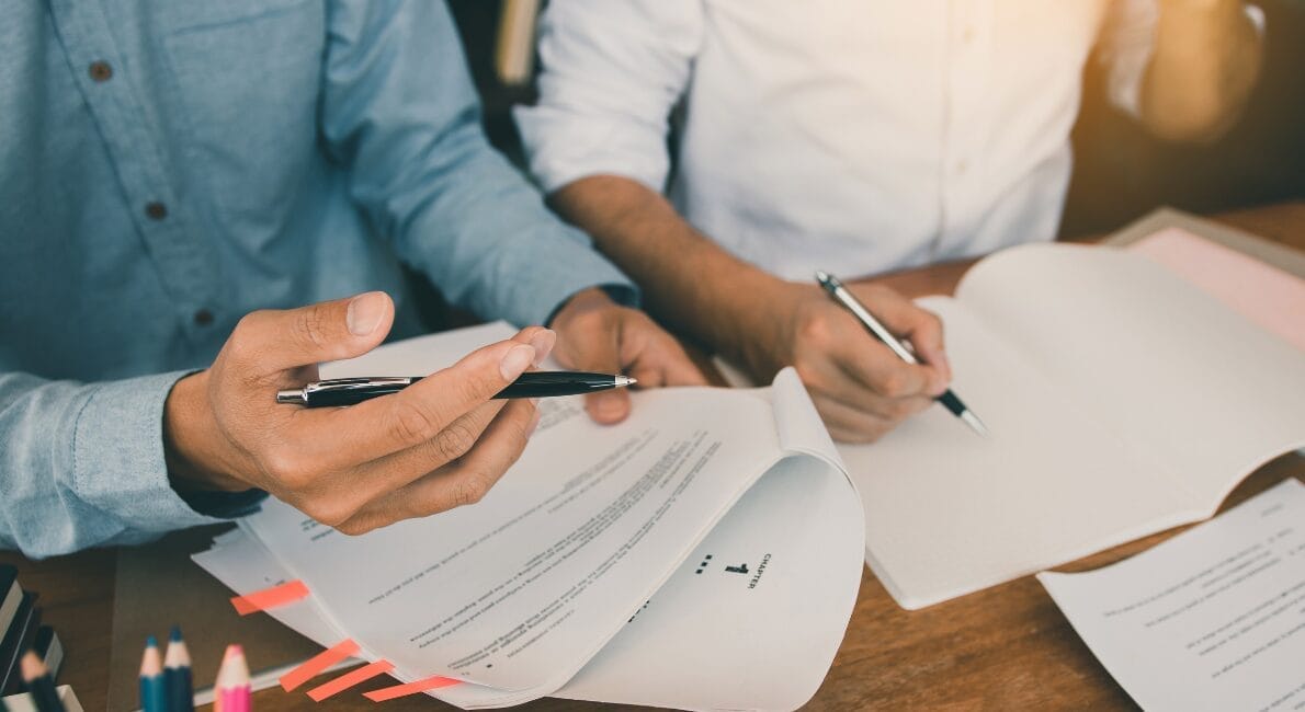 To personer ved et træbord gennemgår dokumenter. Den ene holder en pen og papir med orange klistersedler, den anden skriver på et blankt ark. Sollys oplyser scenen. Forskellige papirer og blyanter ligger spredt på bordet.