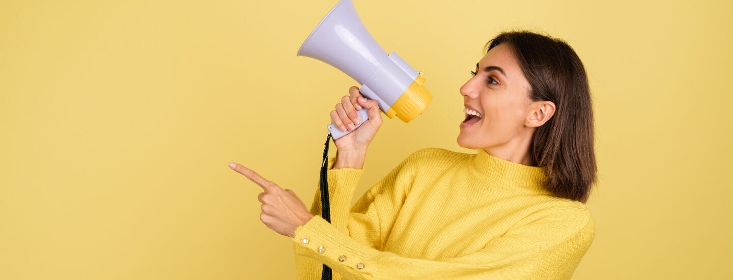 En kvinde i en gul sweater holder en megafon og peger til siden. Hun smiler og står på en solid gul baggrund.