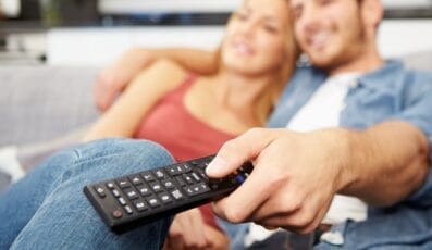 Et par sidder på en sofa, smiler og slapper af. Manden holder en fjernbetjening til fjernsynet og peger den mod fjernsynet. De fremstår komfortable og tilfredse med en hyggelig stue i baggrunden.