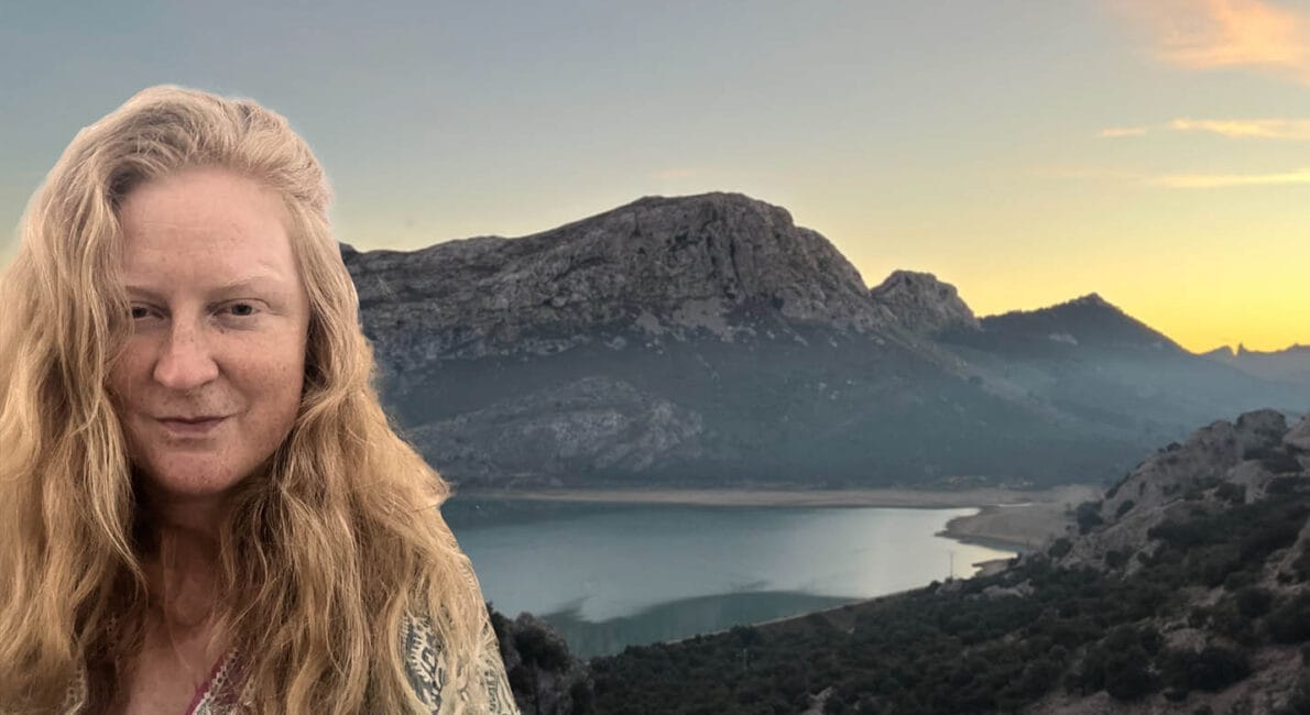 En person med langt blondt hår står foran et naturskønt bjerglandskab ved solnedgang. Bag dem er der et roligt vandområde og forrevne bakker under en farverig himmel, der skifter fra dag til nat.