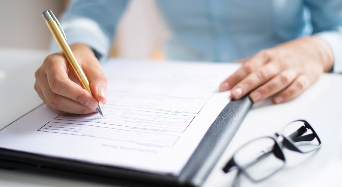 En person iført en lyseblå skjorte udfylder en formular på en udklipsholder med en kuglepen. I nærheden hviler et par sortindfattede briller på en hvid overflade.