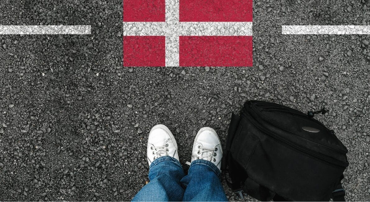 En person iført hvide sneakers og blå jeans står på en grusoverflade ved siden af en sort rygsæk. Foran dem er et rødt rektangel med et hvidt kors, der ligner Danmarks flag, markeret mellem to hvide streger på jorden.