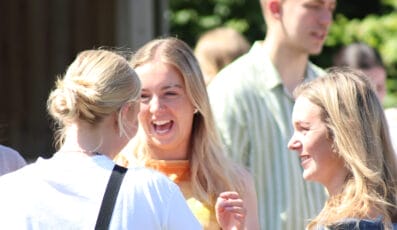 Three people are joyfully conversing outdoors on a sunny day. The central person is laughing, while the others smile. The background is slightly blurred, showing more people and greenery.