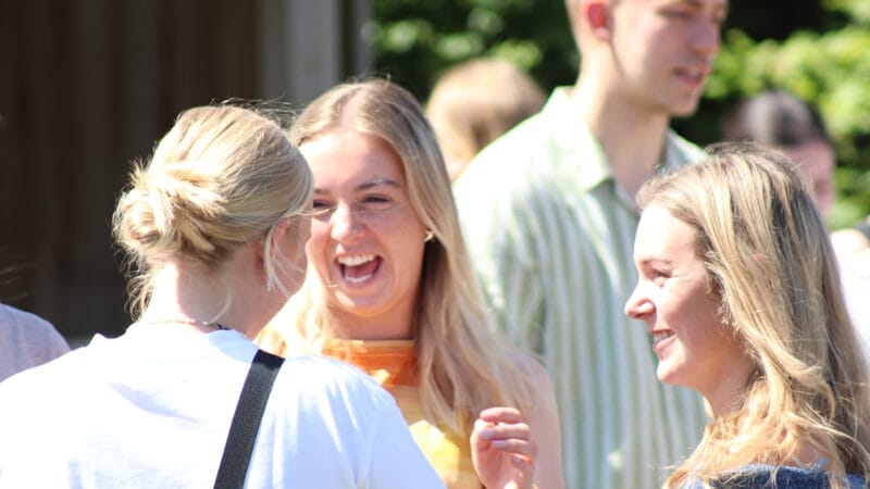 Tre unge er udenfor på en solskinsdag og griner og smiler i samtale. Scenen er livlig med en sløret baggrund af flere mennesker og grønne områder. Fokus er på udtryk for glæde og engagement i gruppen.