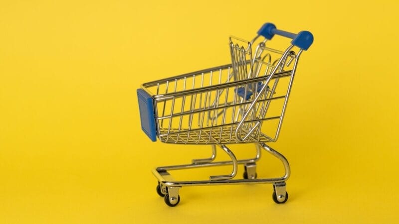 En lille indkøbskurv i metal med blå håndtag er placeret på en solid gul baggrund. Vognen er tom, og dens skinnende overflade afspejler det skarpe lys i billedet.