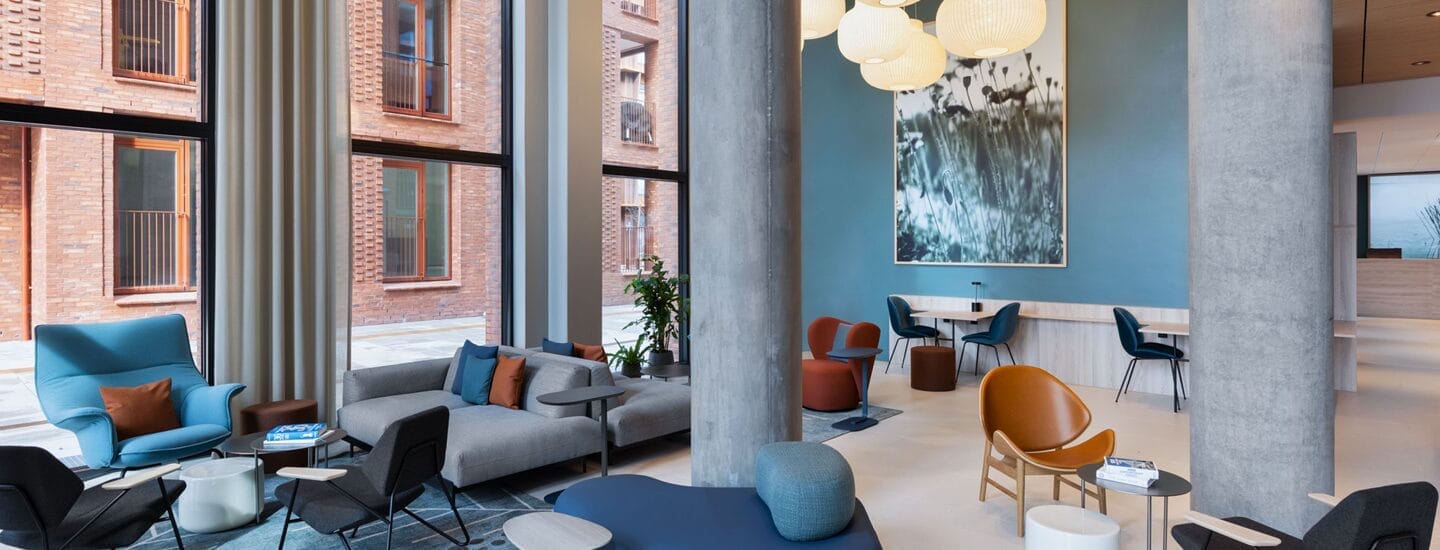 Moderne loungeområde med store vinduer, hyggelige siddepladser i blå og brune farver og en høj betonsøjle. Et stort abstrakt maleri dekorerer den blå væg. Små borde og stole er arrangeret til afslappede siddepladser - perfekt til gæster, der nyder rabat på hotelovernatninger.