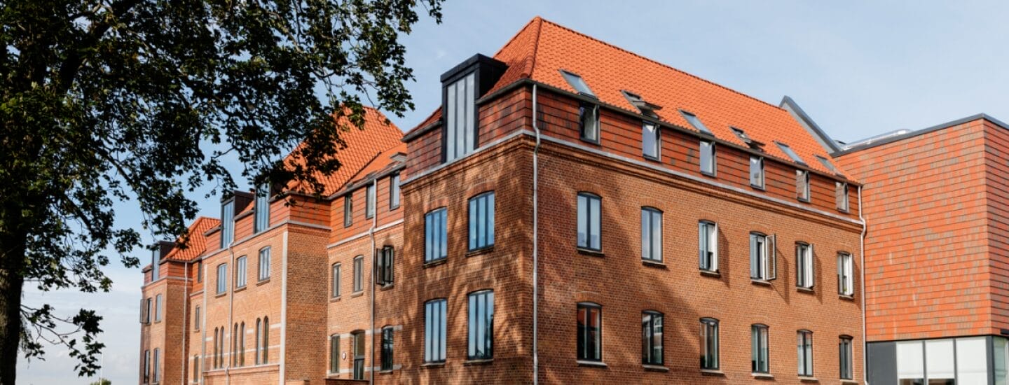 En stor tre-etagers murstensbygning med rødt tegltag og flere vinduer under en blå himmel. Et træ med grønne blade er delvist synligt i venstre side af billedet.