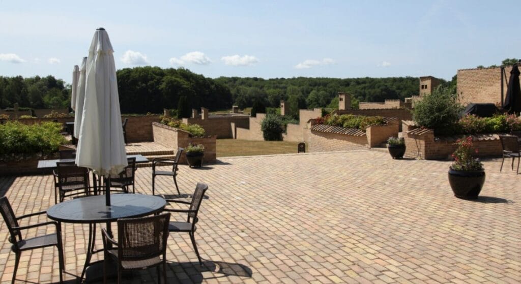 Udendørs terrasse med runde borde, stole og lukkede parasoller på en bred murstensbelagt terrasse. Murstensvægge fremkalder det tidløse design af Utzon fredensborghusene, mens potteplanter og grønt lyser den klare himmel i baggrunden.