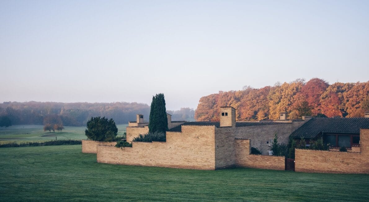 En moderne lys murstensbygning tegnet af Utzon arkitekt står på en græsplæne. Baggrunden viser en række træer med efterårsløv under en klar himmel.